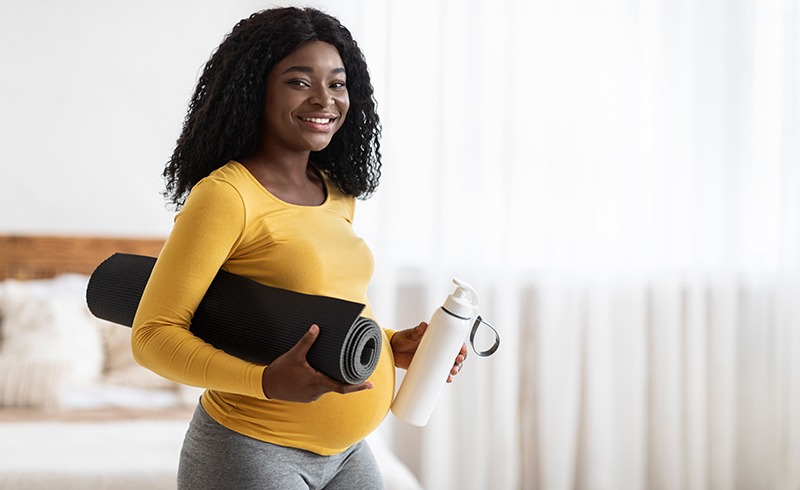 Happy black pregnant woman with yoga mat and bottle of water looking at camera and smiling. 