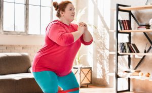 Young obese woman doing squats with a fitness band.